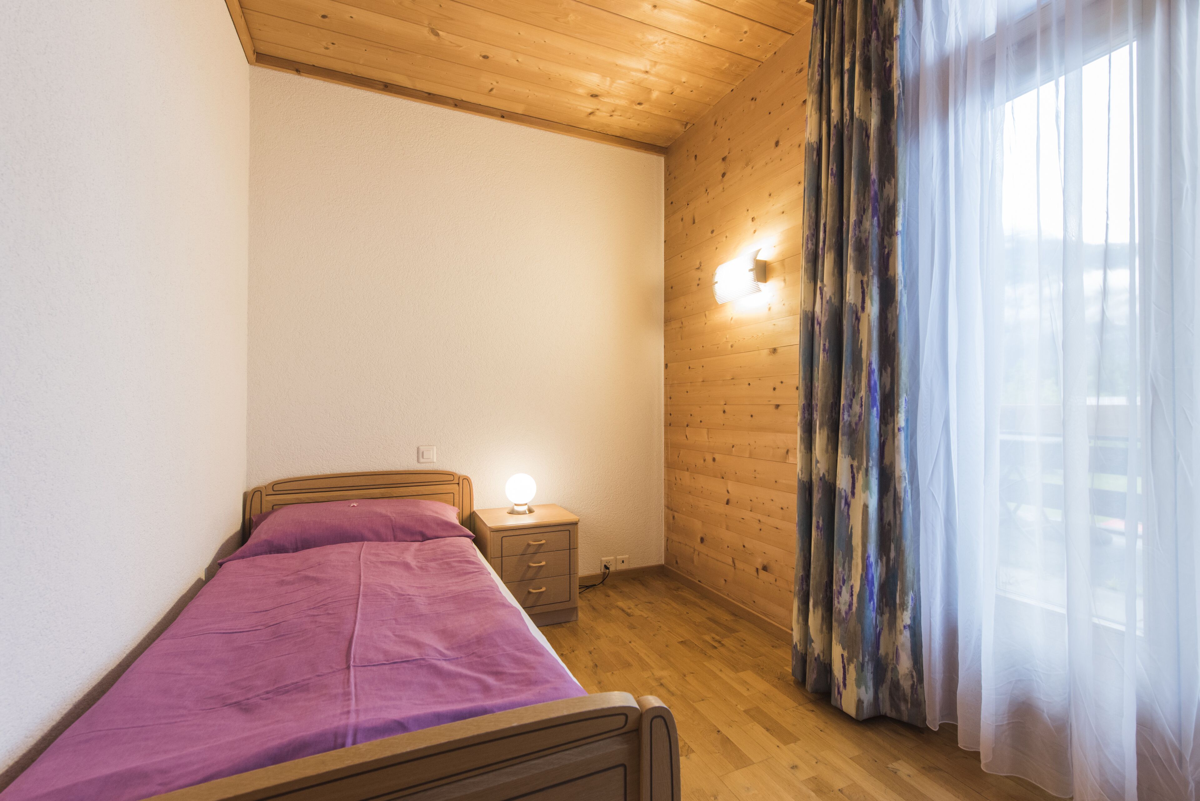 A bedroom with a wooden bed, a nightstand with a lamp, a wooden wall, and a window with sheer curtains.
