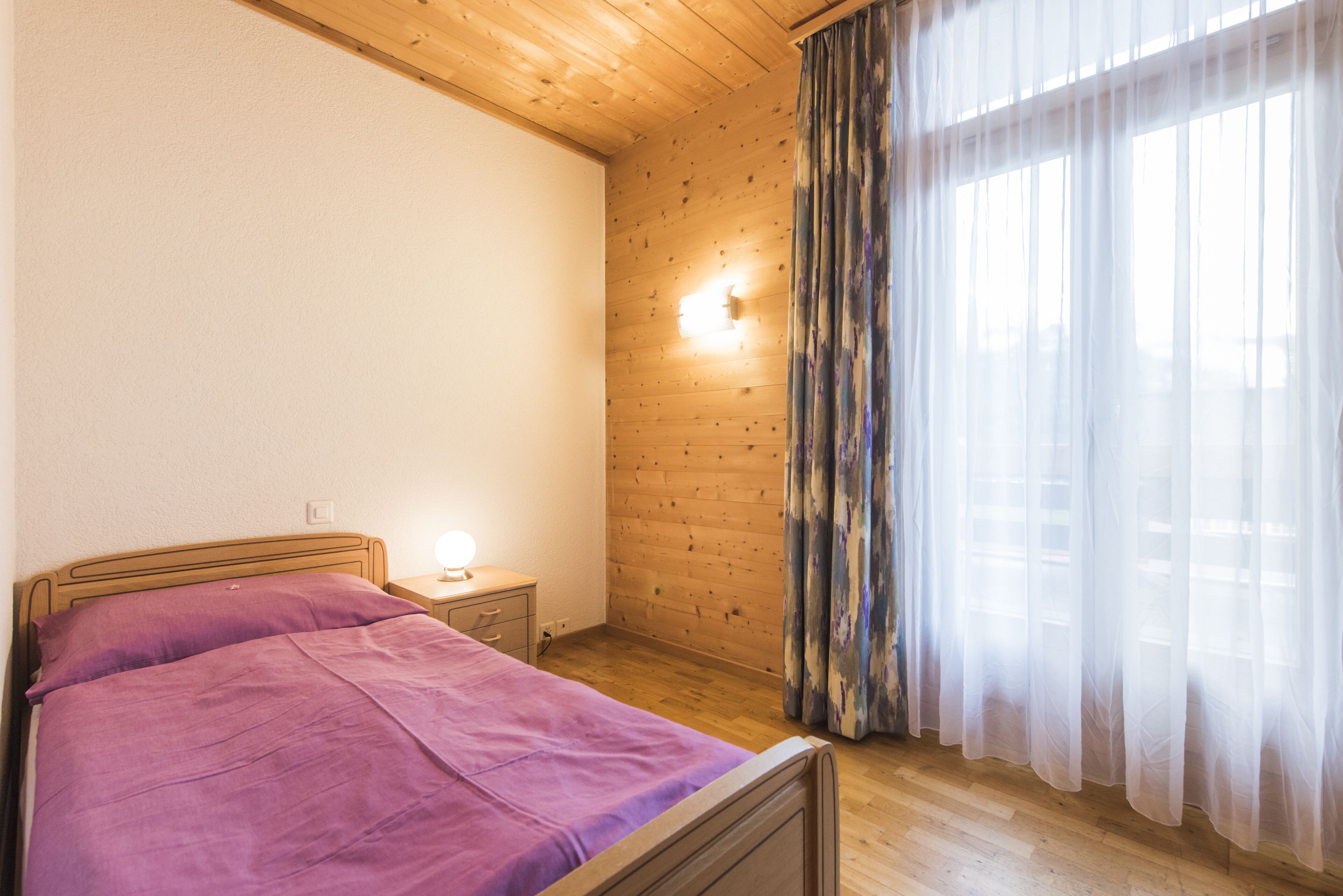 A bedroom with a wooden ceiling, a bed with a purple bedspread, a nightstand with a lamp, and a window with sheer curtains.
