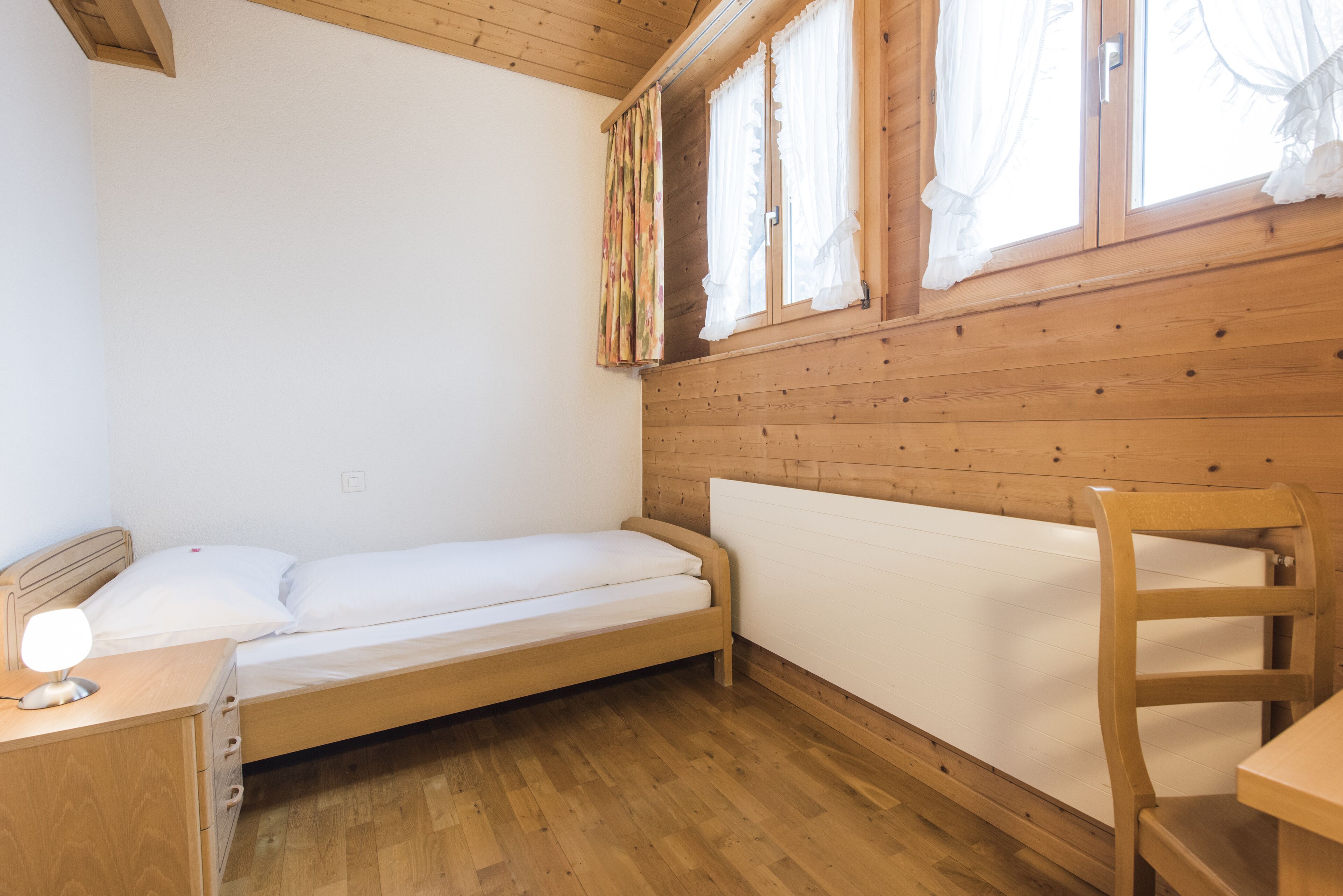 A single bed with a wooden headboard, a bedside table with a lamp, and a window with curtains.