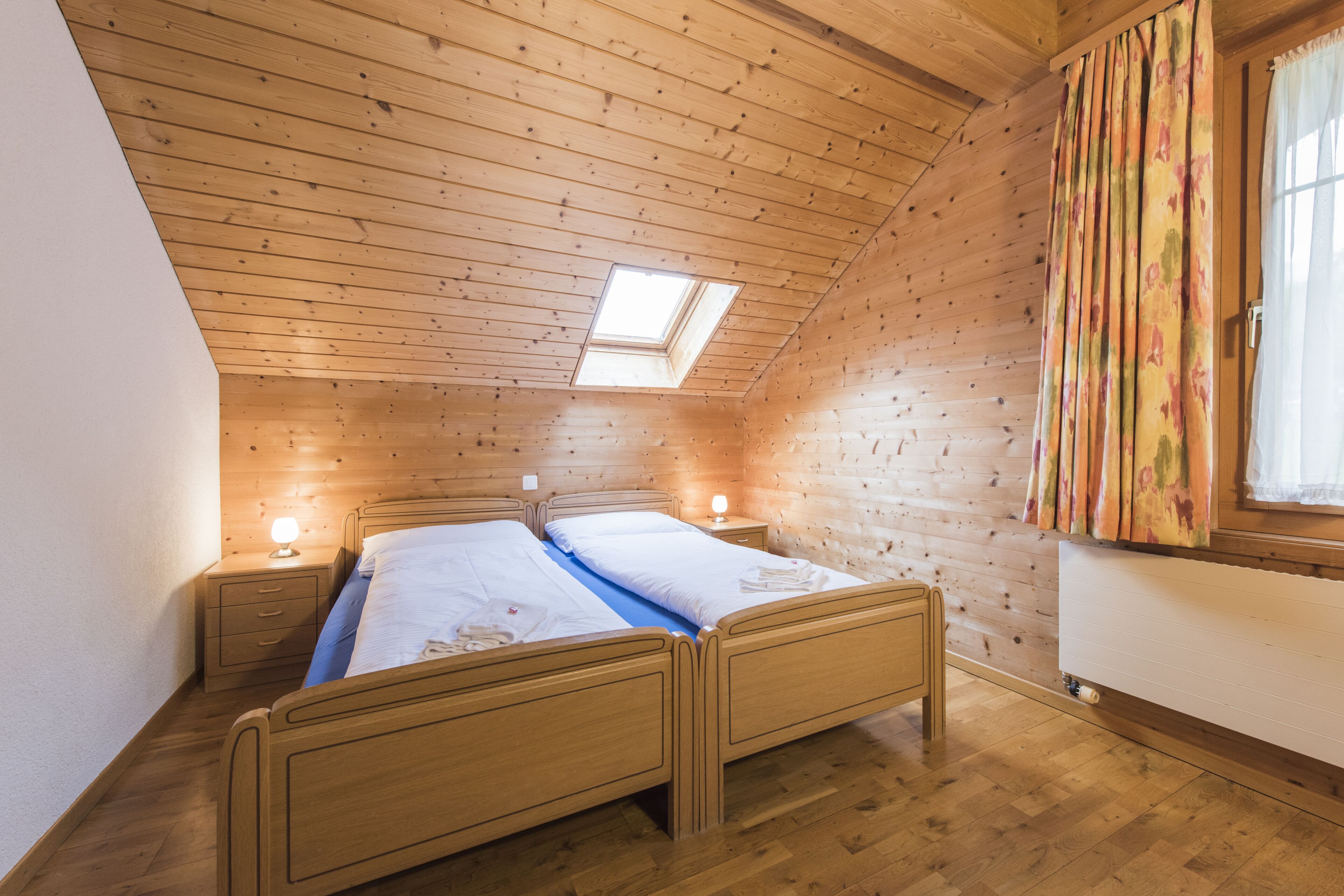 Ein gemütliches Schlafzimmer mit Holzbalkendecke, Dachfenster, einem Bett mit zwei Nachttischen und einem Fenster mit Vorhängen.