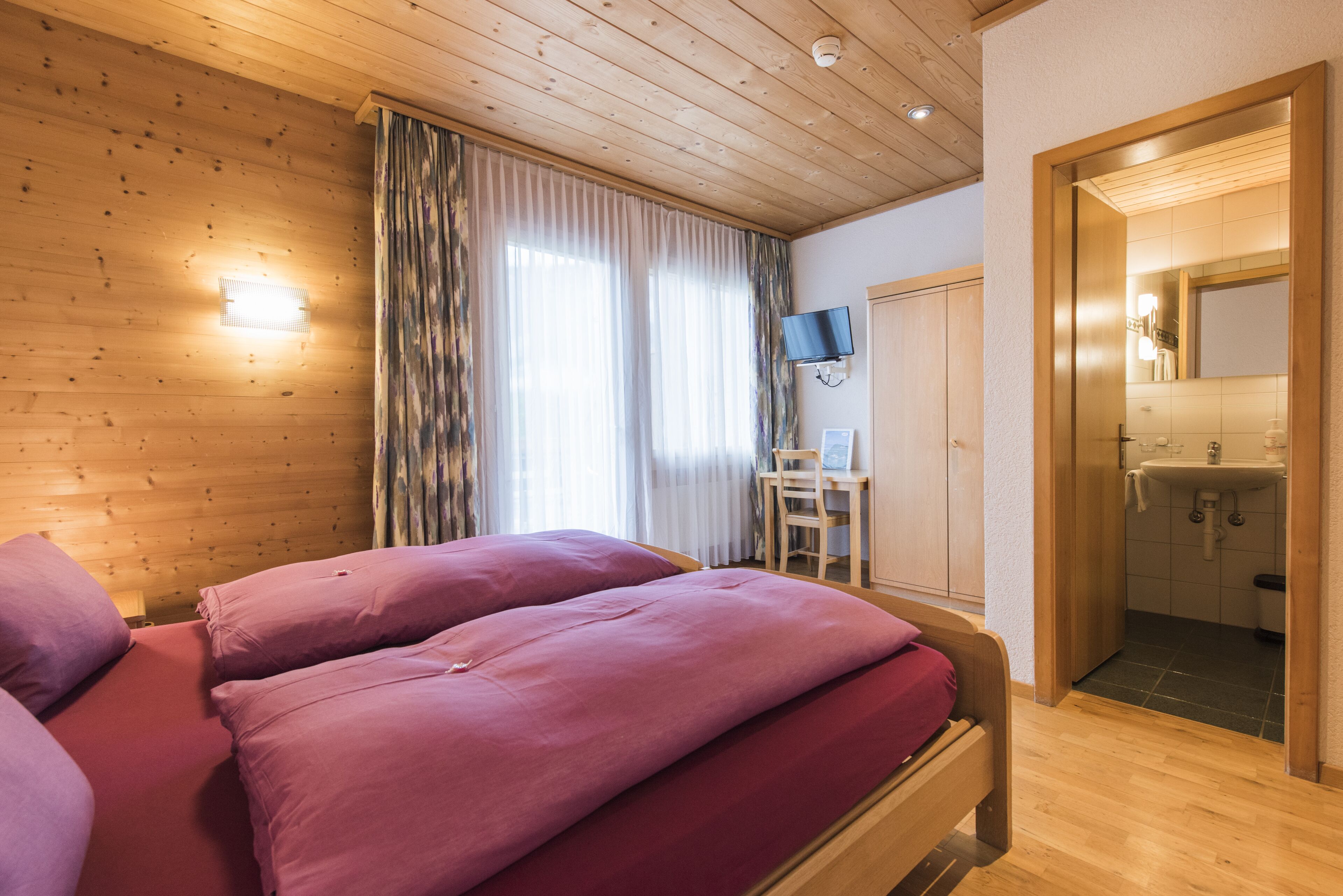 Una habitación con paneles de madera, una cama, un escritorio, un televisor y un baño visible a través de una puerta abierta.