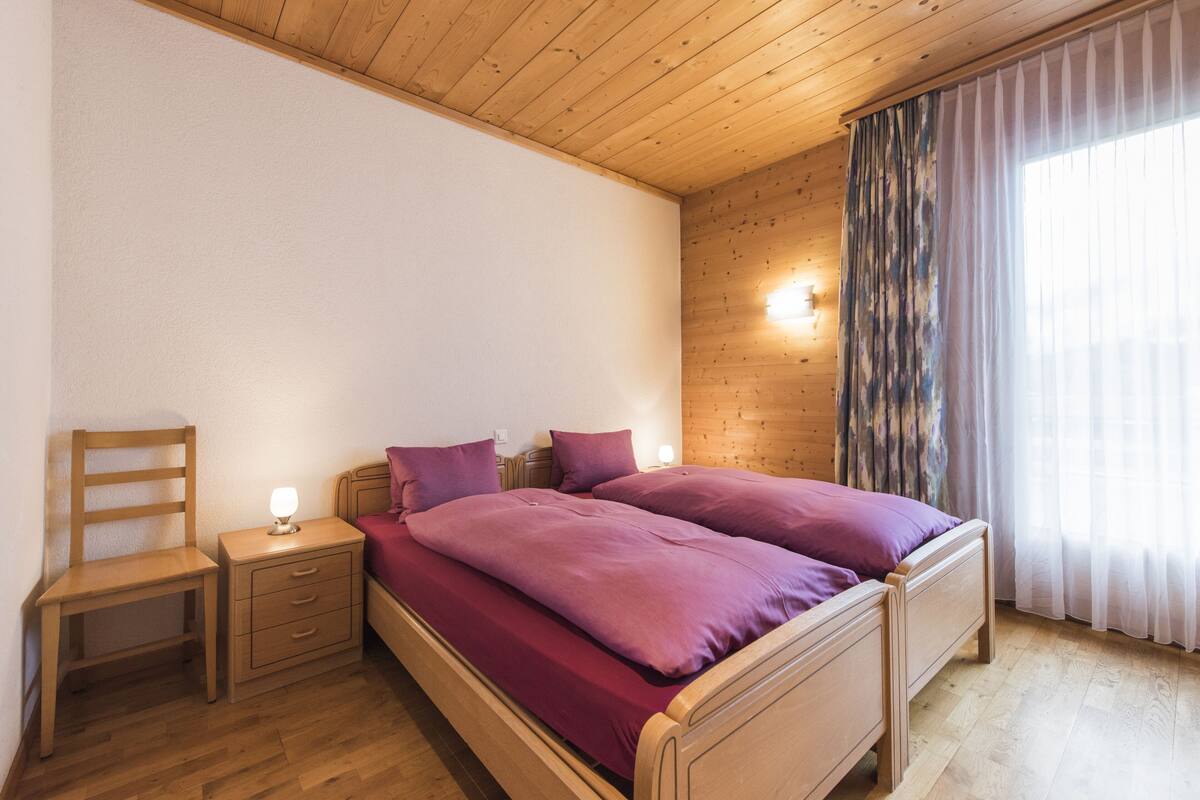 A bedroom with a wooden bed, a nightstand, a chair, and a window with curtains.