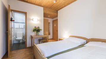 Ein Hotelzimmer mit Holzboden, einem Schreibtisch mit Stuhl, einem Bett mit zwei Kissen und einem durchgehenden Türspalt einsehbaren Badezimmer.