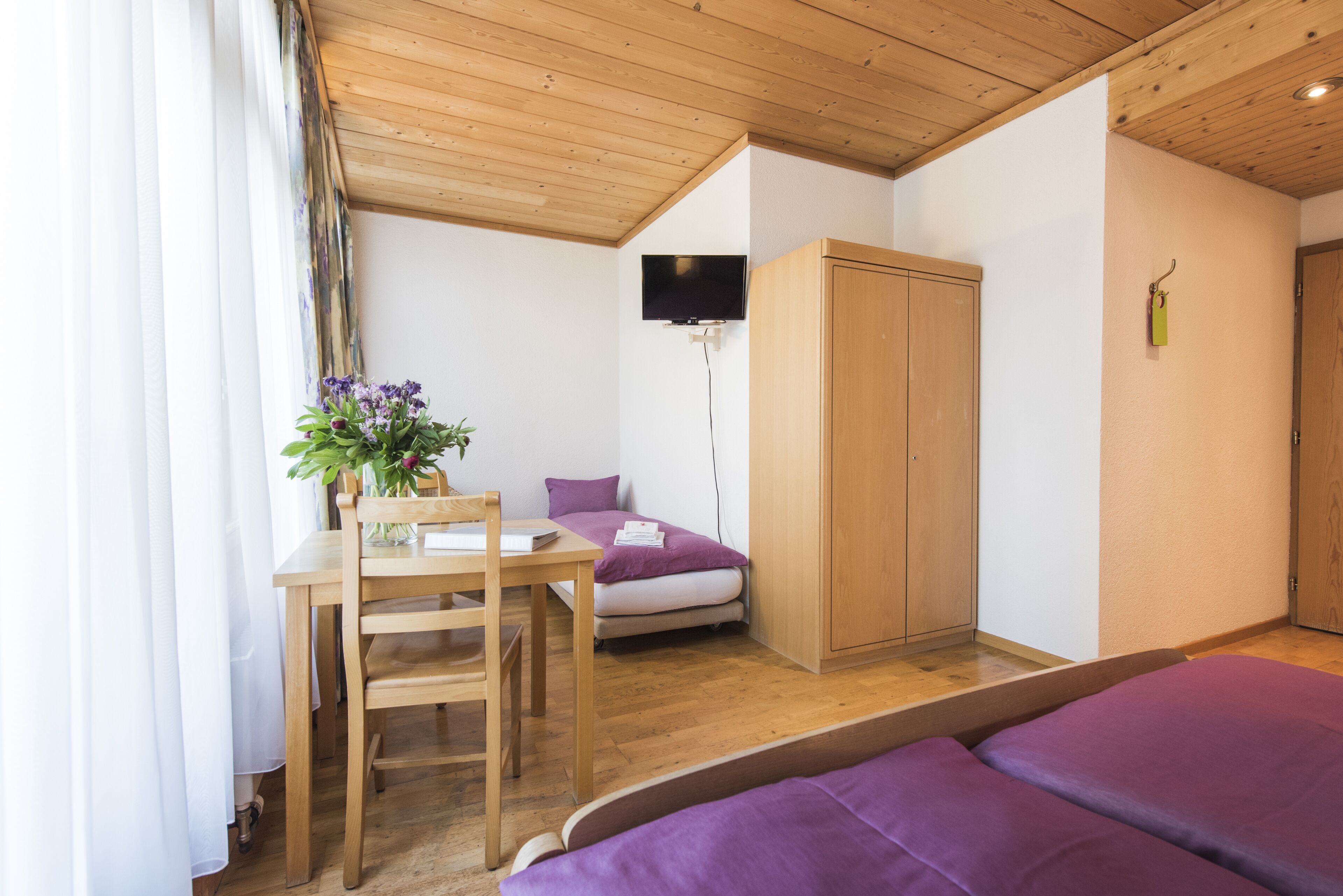 Une chambre avec un plafond en bois, un lit avec une literie violette, une armoire en bois, un téléviseur fixé au mur et une petite table avec une chaise et un vase de fleurs.