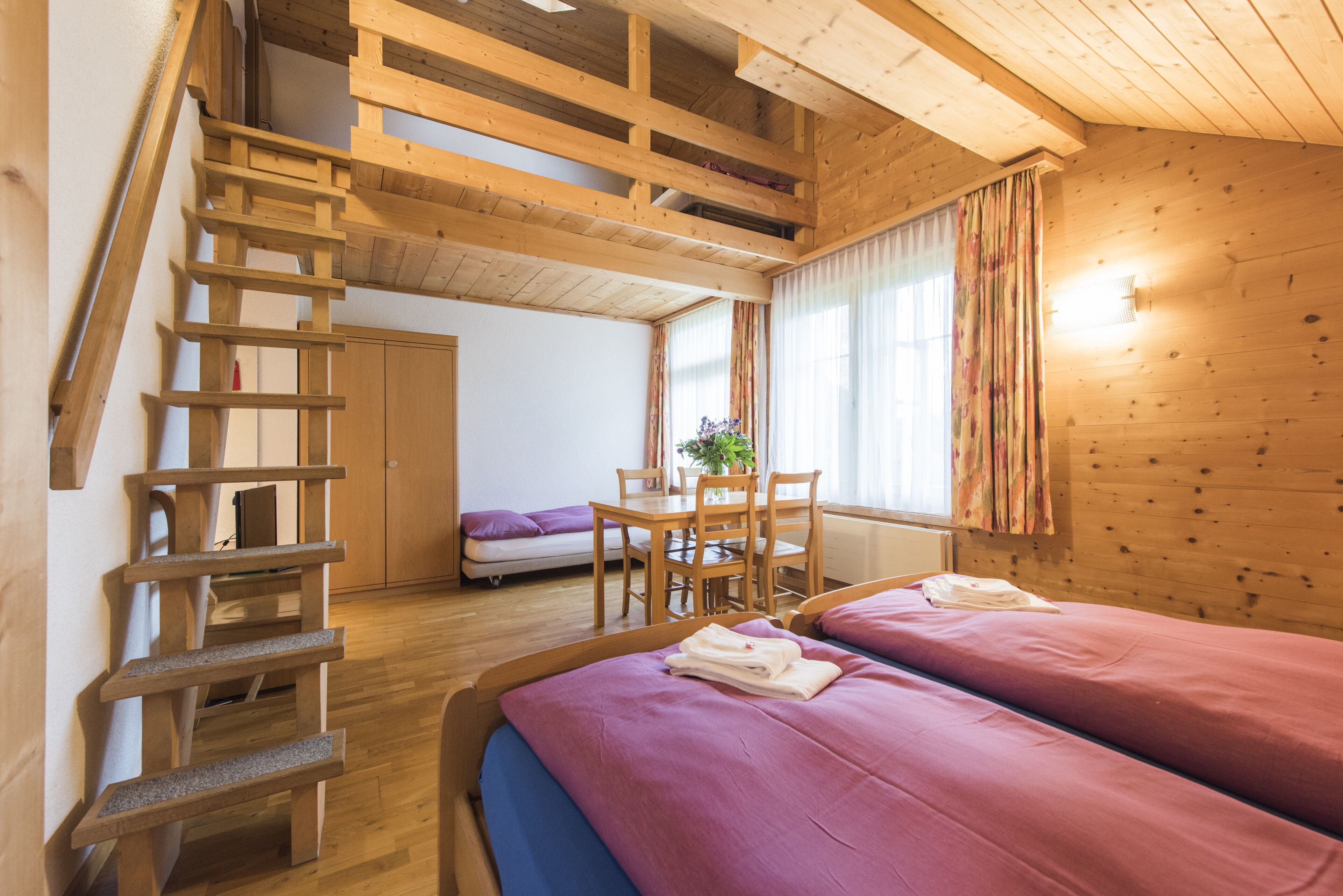 Un interior de madera con una escalera, dos camas con ropa de cama rosa, un comedor y un ventanal con cortinas.