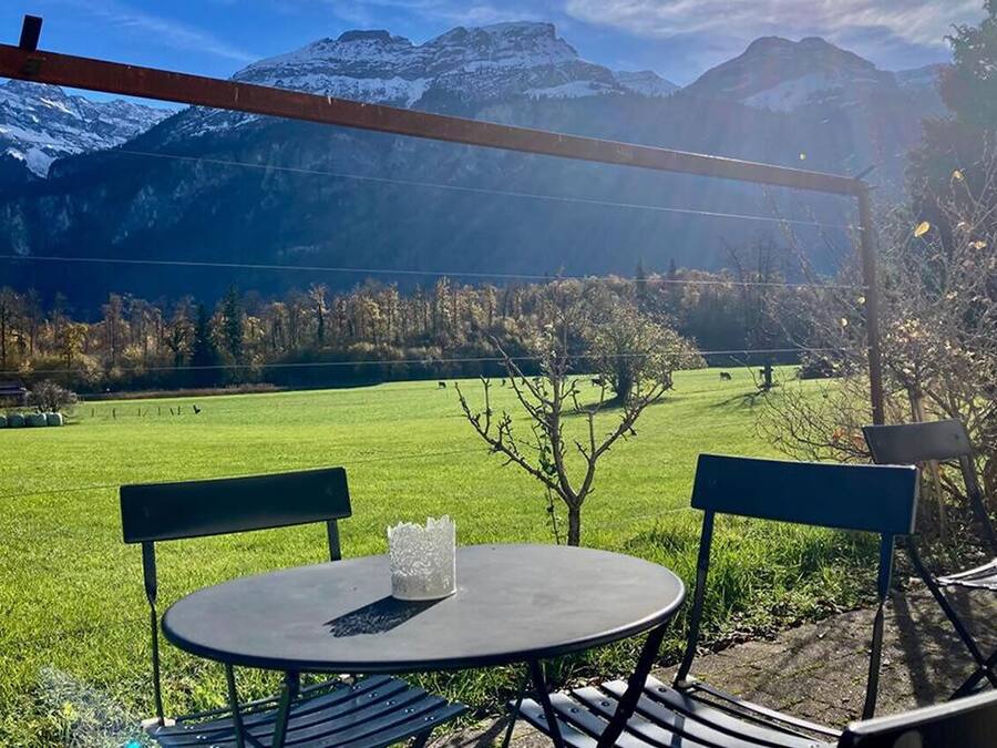 An outdoor seating area with a table and chairs, overlooking a green field and mountains.
