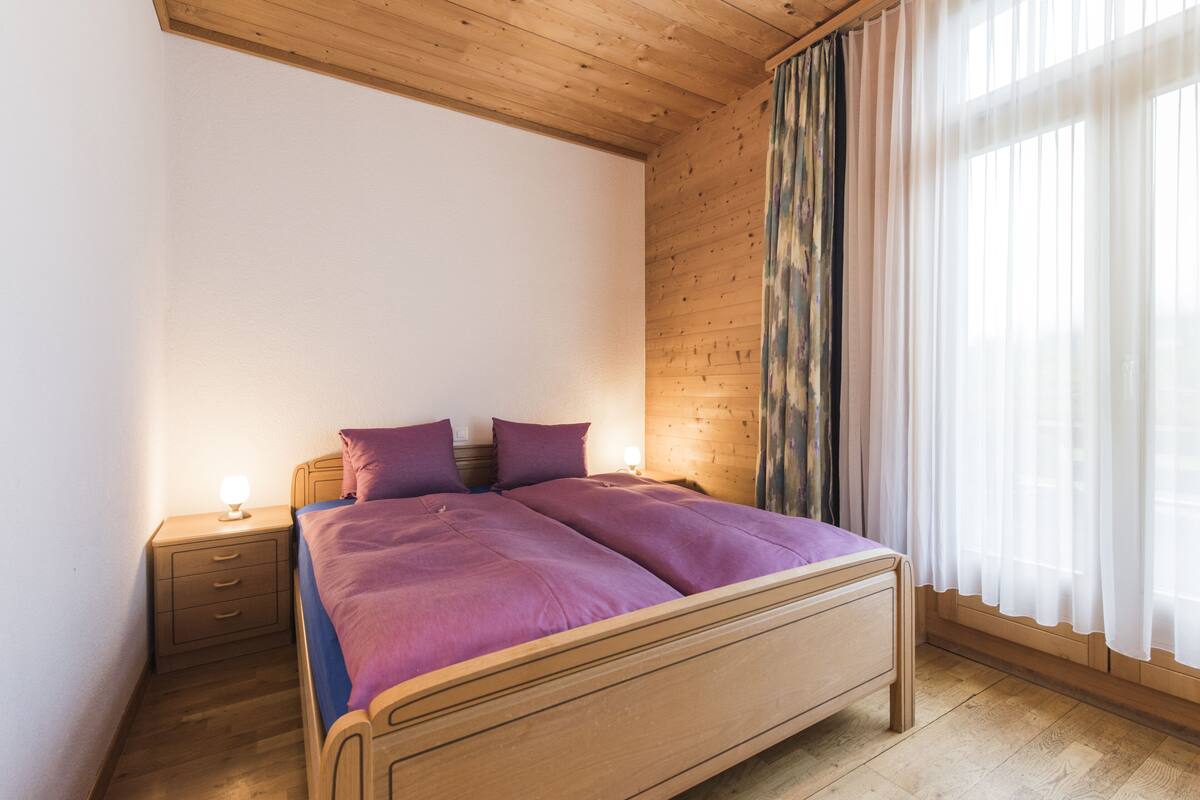A bedroom with a wooden bed, a nightstand, and a window with sheer curtains.