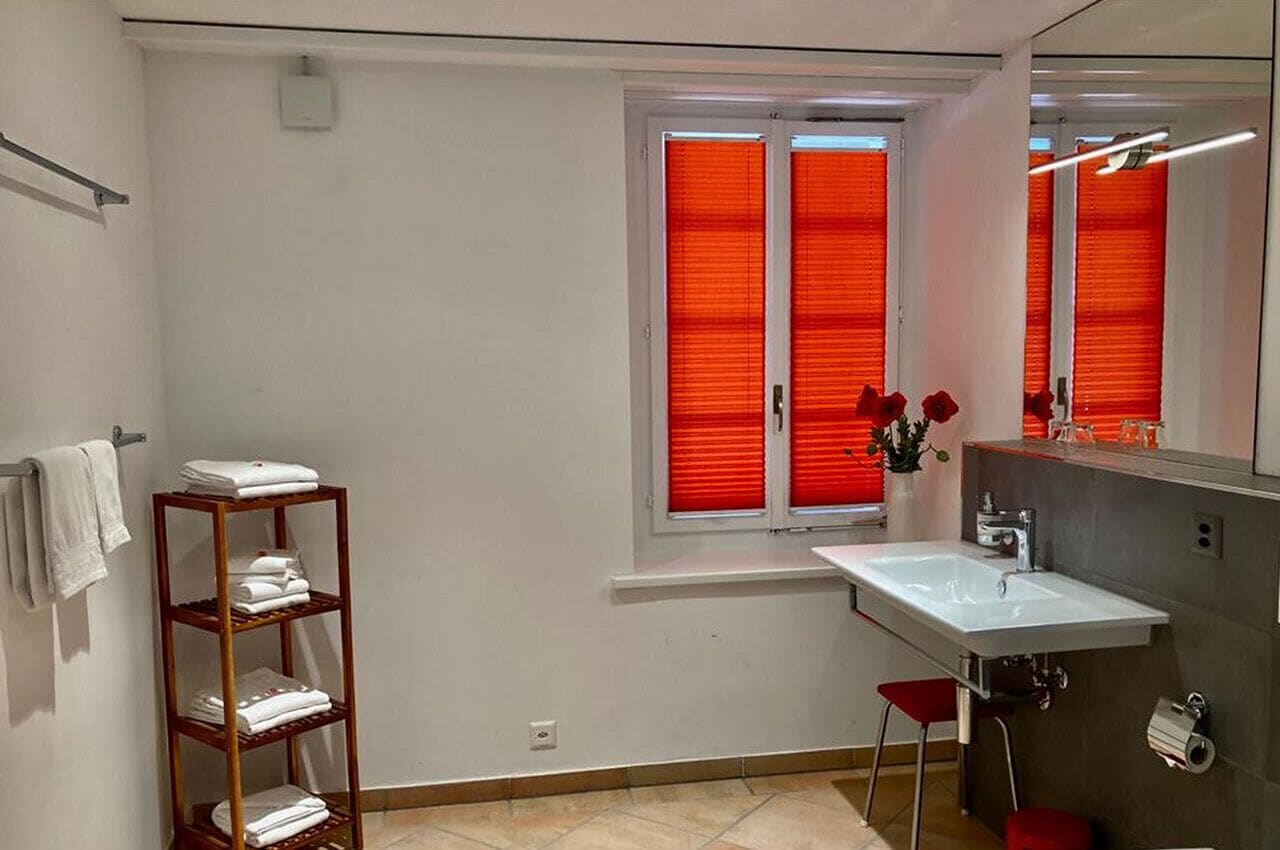 Un baño con un lavabo blanco, una estantería de madera con toallas, un ventanal rojo y un espejo.