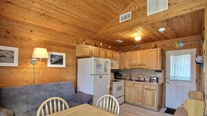 A cozy cabin interior with a wooden ceiling, a kitchenette, a dining area with a table and chairs, and a sofa.