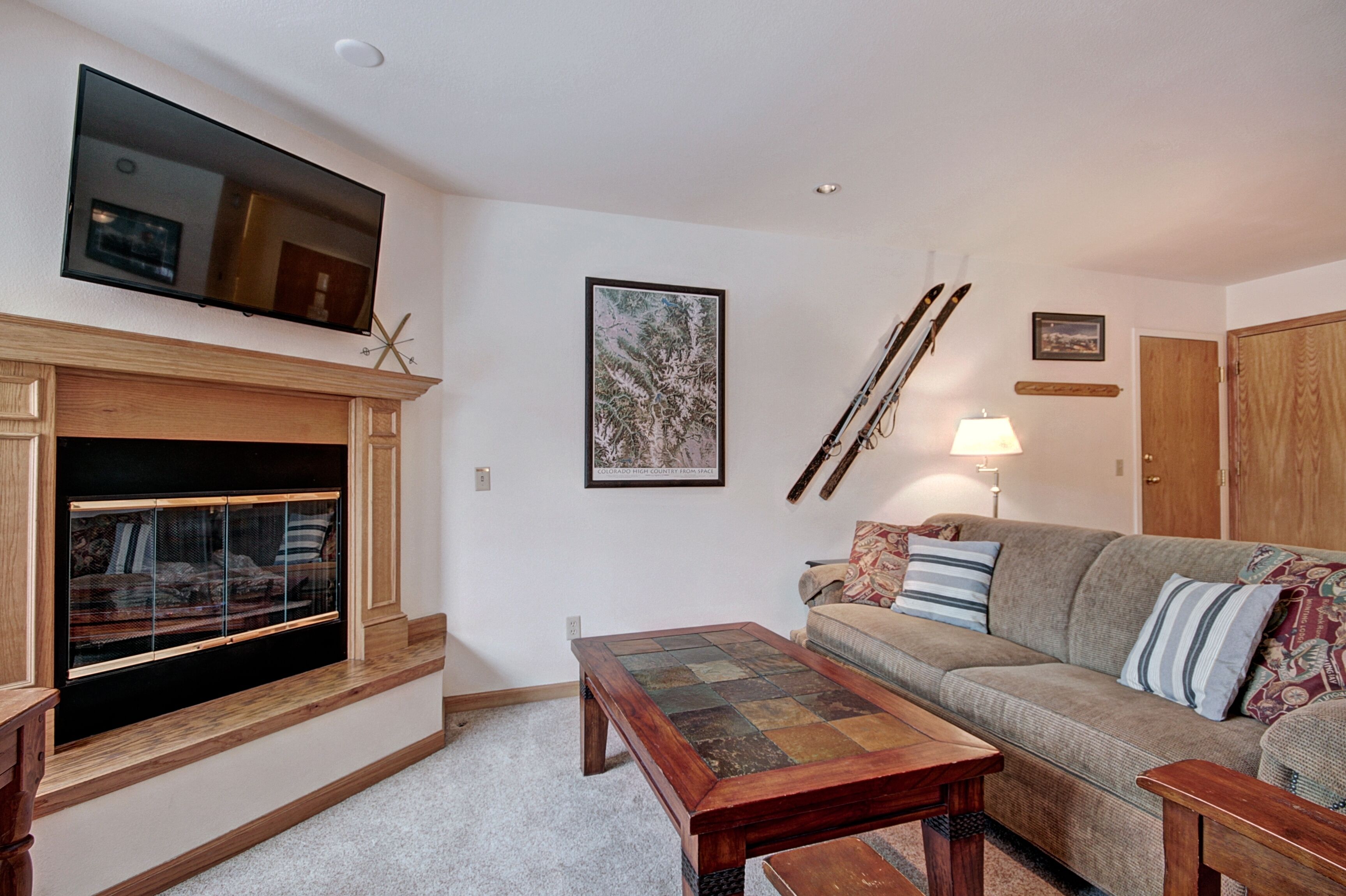 A living room with a fireplace, a television mounted above it, a sofa with cushions, a coffee table, and skis leaning against the wall.