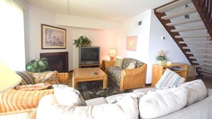 A living room with a sofa, a coffee table, a TV, a lamp, and a staircase. - Crab Cove On The Bay 406 (Ocean City)