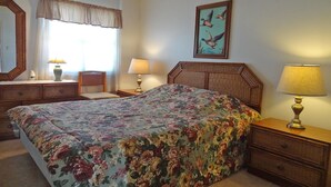 Une chambre à coucher avec une couvre-lit à motifs floraux, une tête de lit en bois, une table de chevet avec une lampe, une chaise, une commode et un tableau représentant des canards accroché au mur.