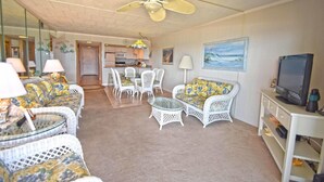A living room with wicker furniture, a ceiling fan, a TV, and a painting of a beach scene. - Ocean Hideaway 503 (Ocean City)
