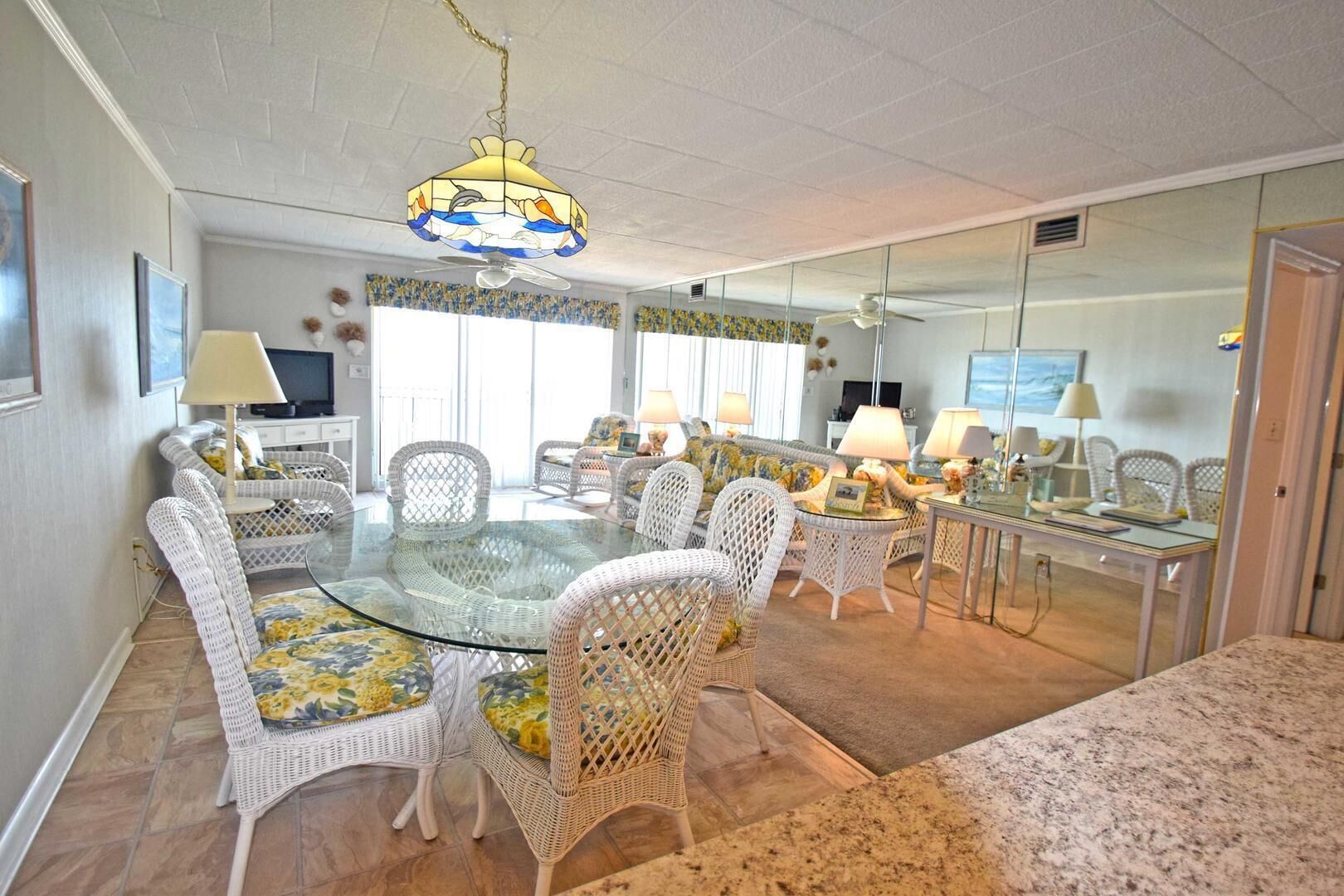 A dining area with wicker chairs and a glass table, a ceiling fan, and a chandelier.