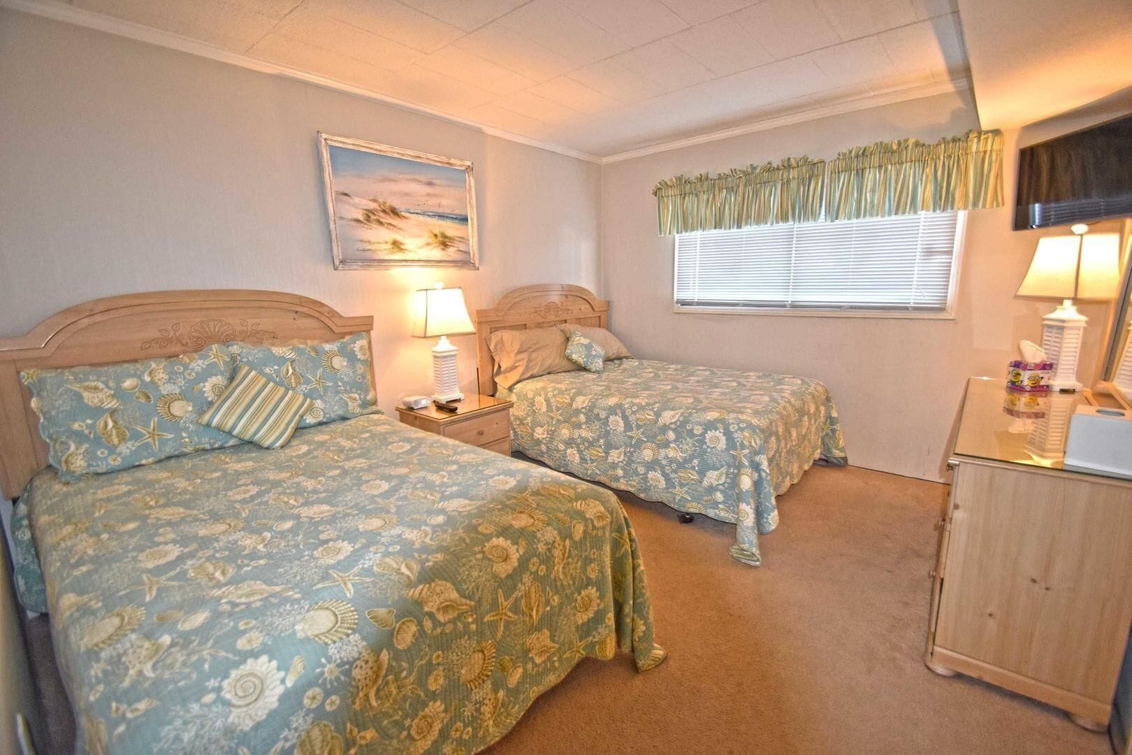 A bedroom with two beds, each with floral bedding, a nightstand, a lamp, and a painting on the wall.
