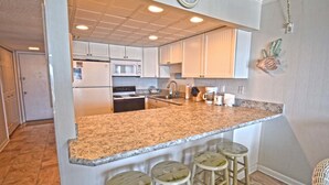 A kitchen with white cabinets, a granite countertop, a refrigerator, a stove, and a microwave. There are three stools at the counter. - Ocean Hideaway 503 (Ocean City)