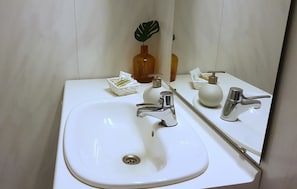 A bathroom with a white sink, two faucets, a mirror, a soap dispenser, and a small plant. - Blåtind Apartments (Fjord)