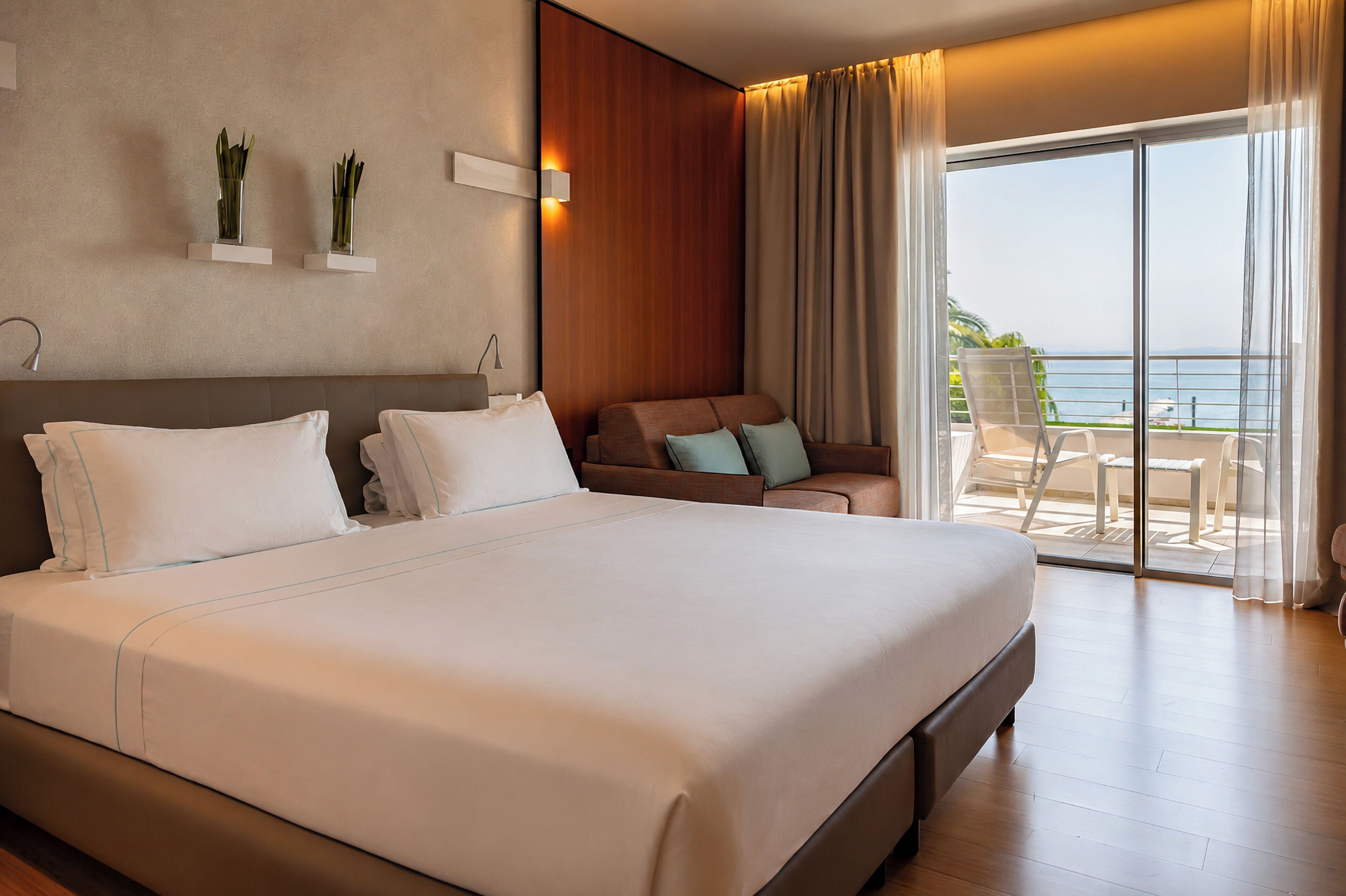 A hotel room with a large bed, a sofa, a balcony with a view of the sea, and a wall-mounted plant.