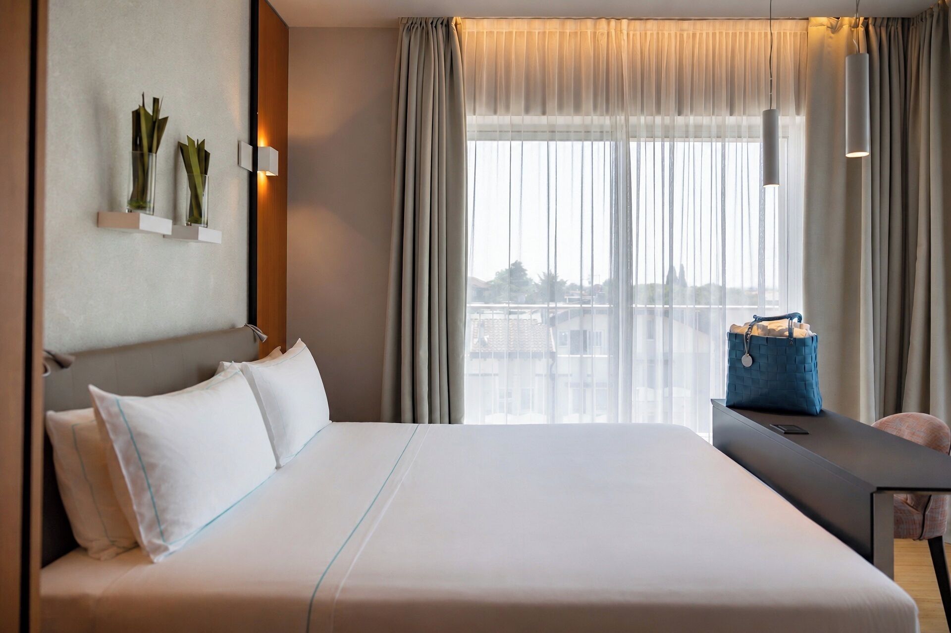 A hotel room with a large bed, a bedside table, a lamp, a window with curtains, and a view of buildings.