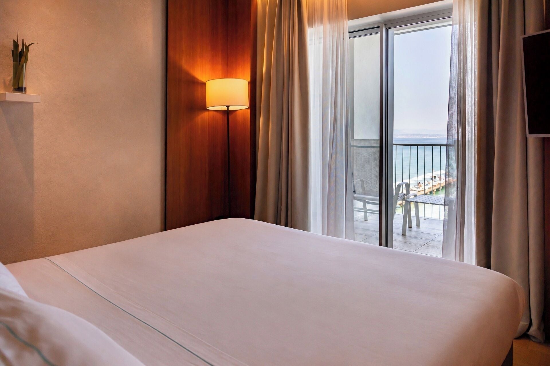 Habitación de hotel con cama, lámpara de pared, vista al balcón y televisión en la pared.