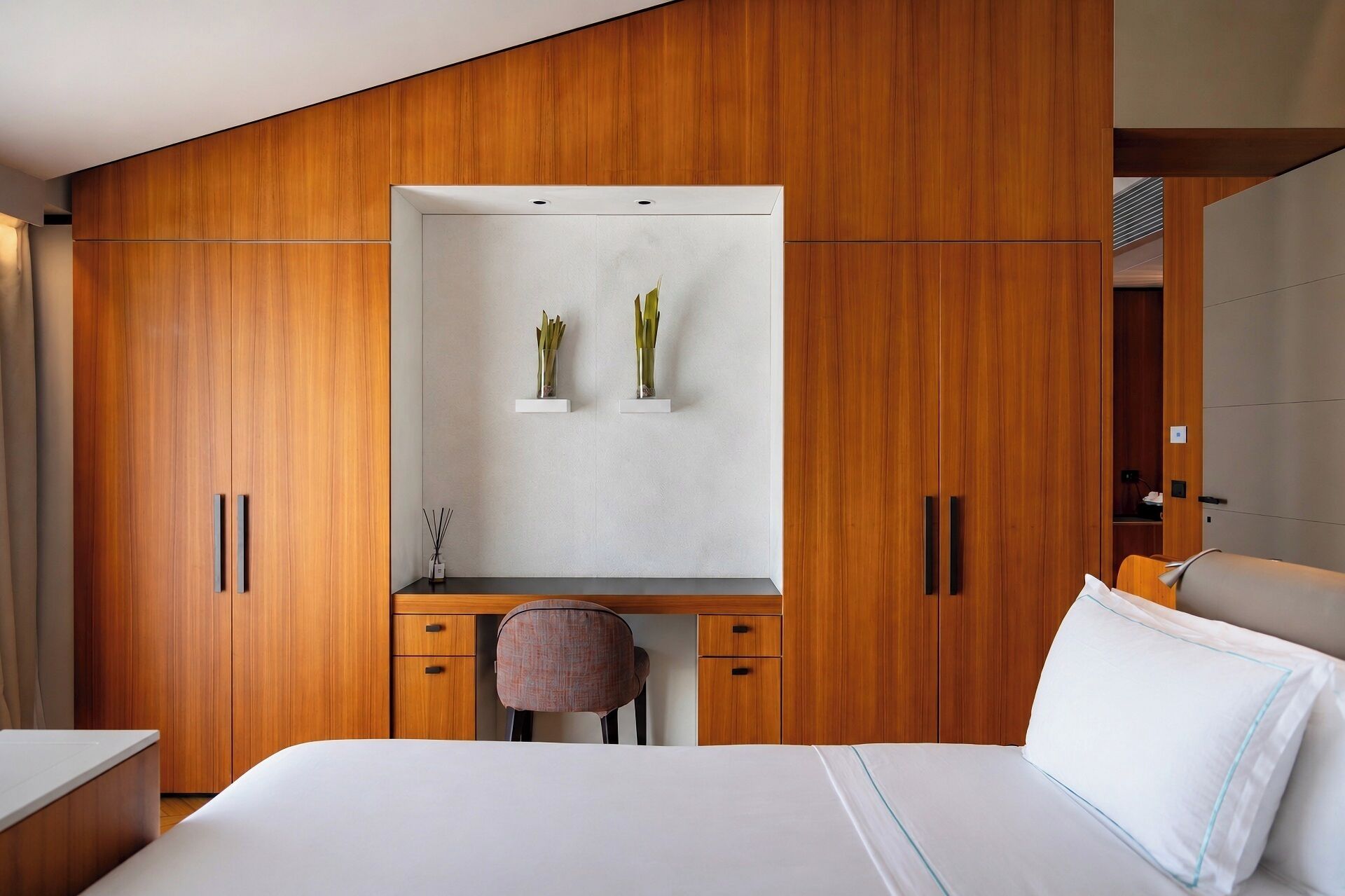 A bedroom with a bed, a desk, and a chair. There are wooden cabinets and a mirror.