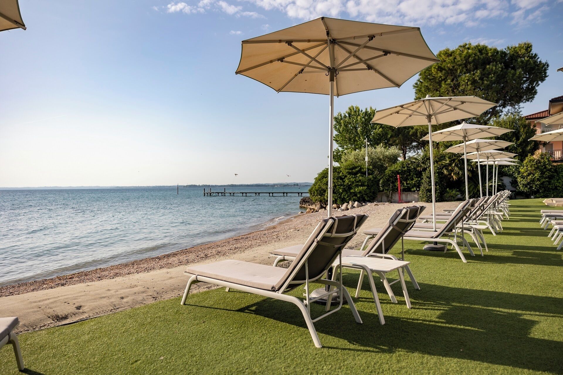 A beachside lounge area with sun loungers and umbrellas, a clear view of the sea, and a wooden pier in the background.