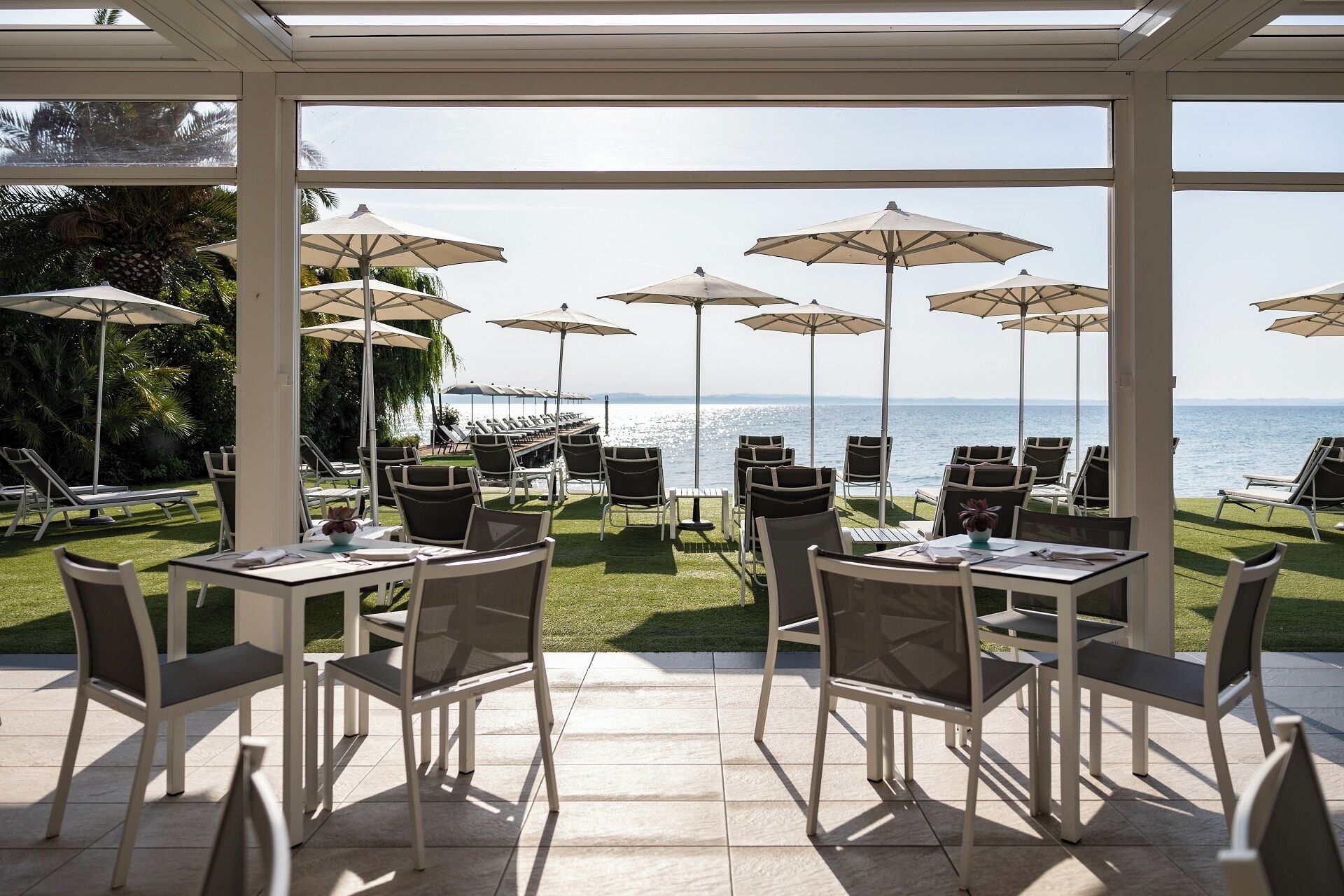 Un área de comedor al aire libre iluminada por el sol, con mesas, sillas y sombrillas, y vista al mar.
