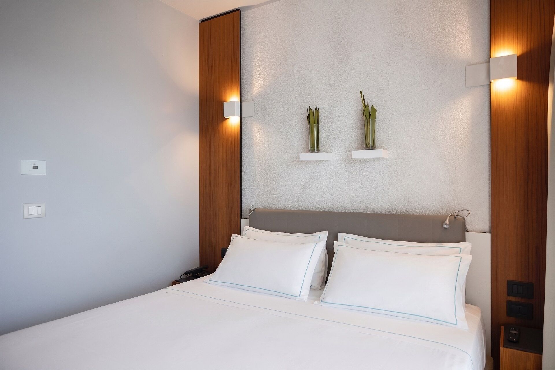 A hotel room with a bed, bedside controls, a wall-mounted light, and two small shelves with decorative items.