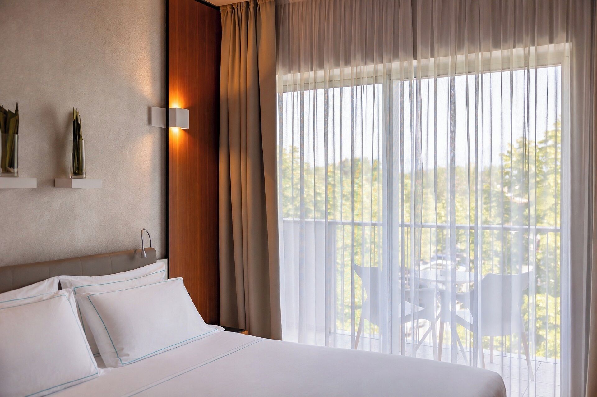 Een hotelkamer met een bed, nachtkastje, een wandlamp en een raam met dunne gordijnen.