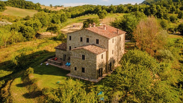 Un bâtiment en pierre, surmonté d’un toit en tuiles, entouré d’une végétation luxuriante et de collines.