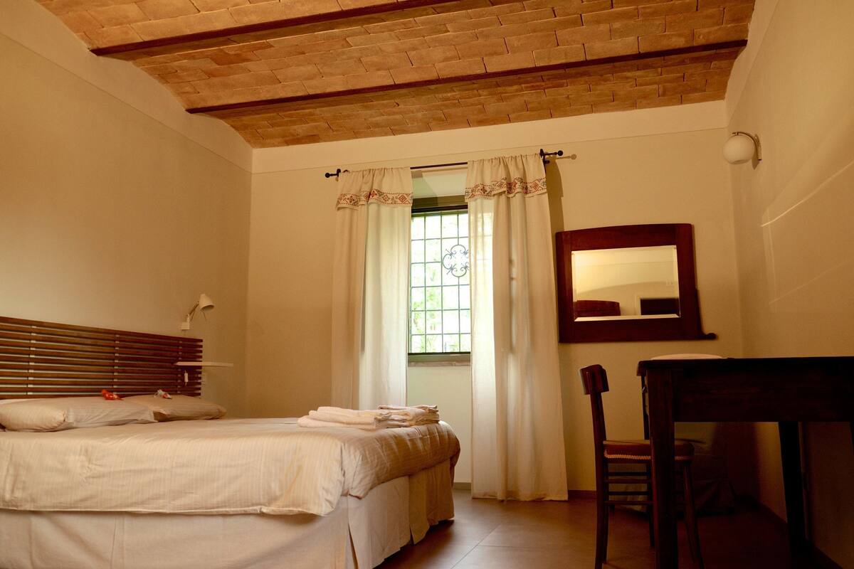 Un dormitorio con techo de madera, una cama con ropa blanca, una ventana con cortinas, una silla y un escritorio.