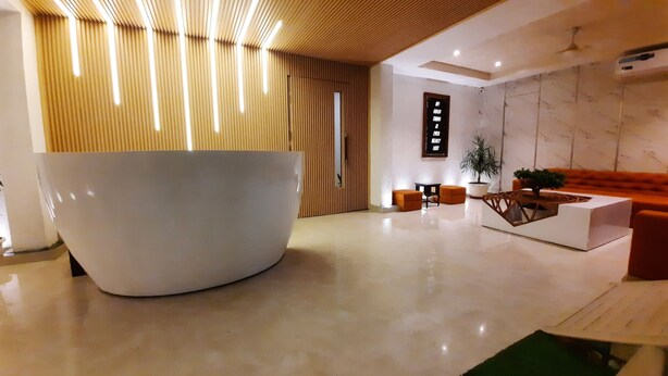 A modern lobby with a large white reception desk, wooden slatted walls, a potted plant, and a seating area with orange chairs. - Hotel Sai Village Gurgaon (Gurugram)