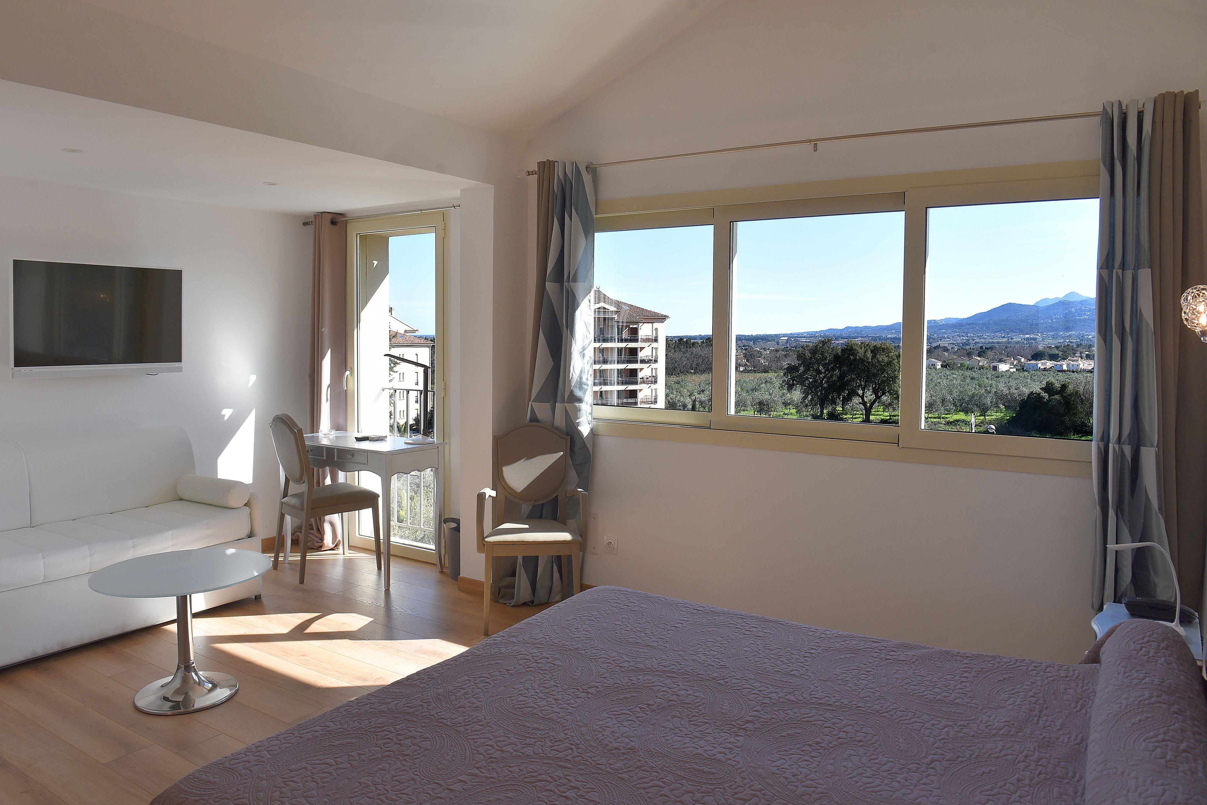 A bedroom with a large bed, a TV mounted on the wall, a dining area with a table and chairs, and a view of the outdoors through a window.