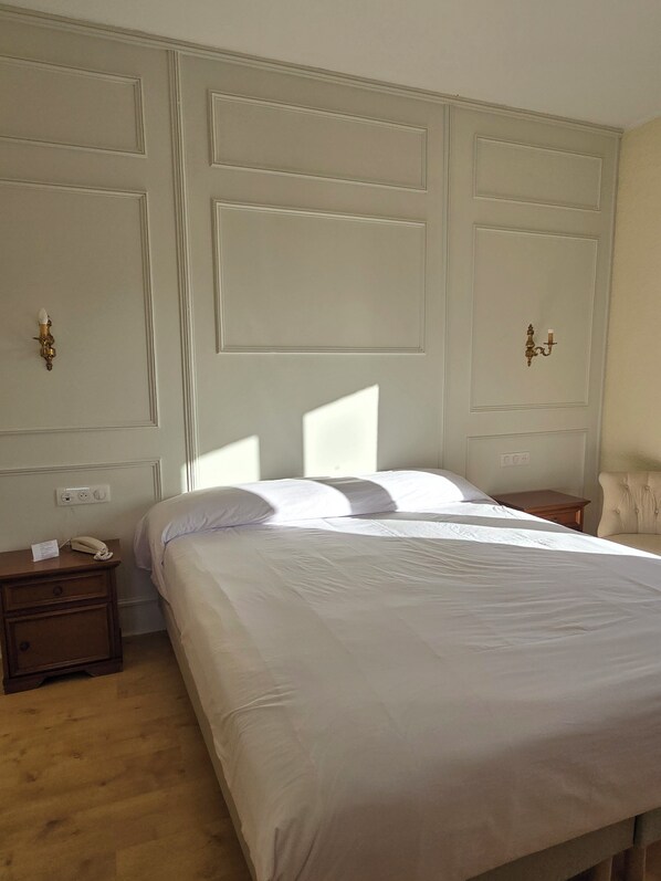 A neatly made bed in a room with a large, plain wall and a small nightstand with a phone and a lamp.
