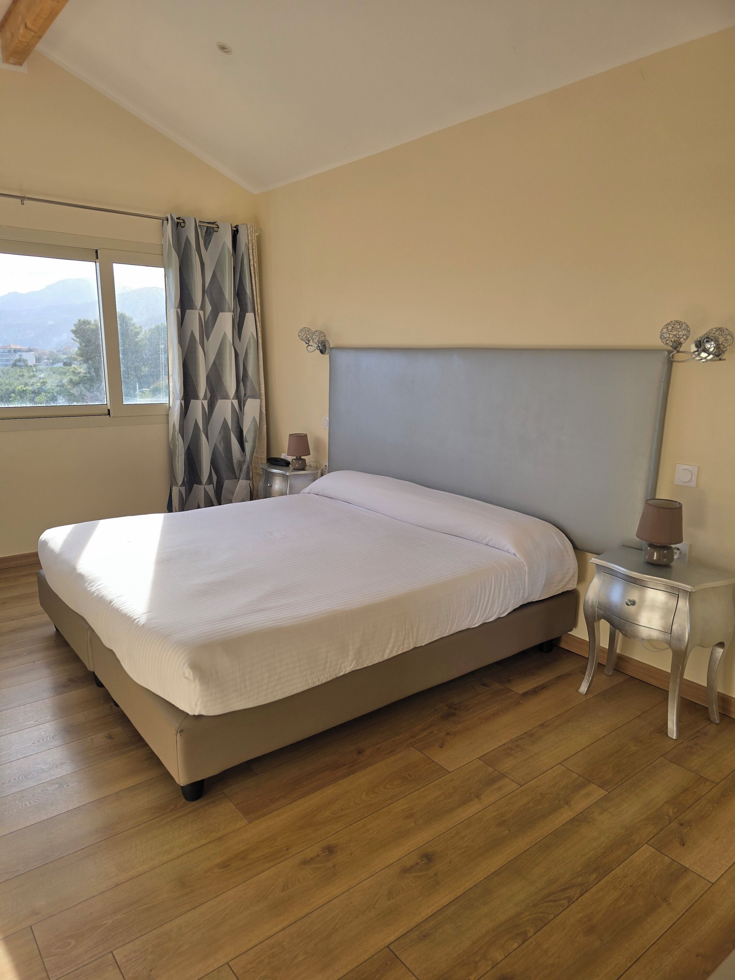 Une chambre à coucher avec un grand lit, un plancher en bois, une table de chevet avec une lampe et une fenêtre avec des rideaux.