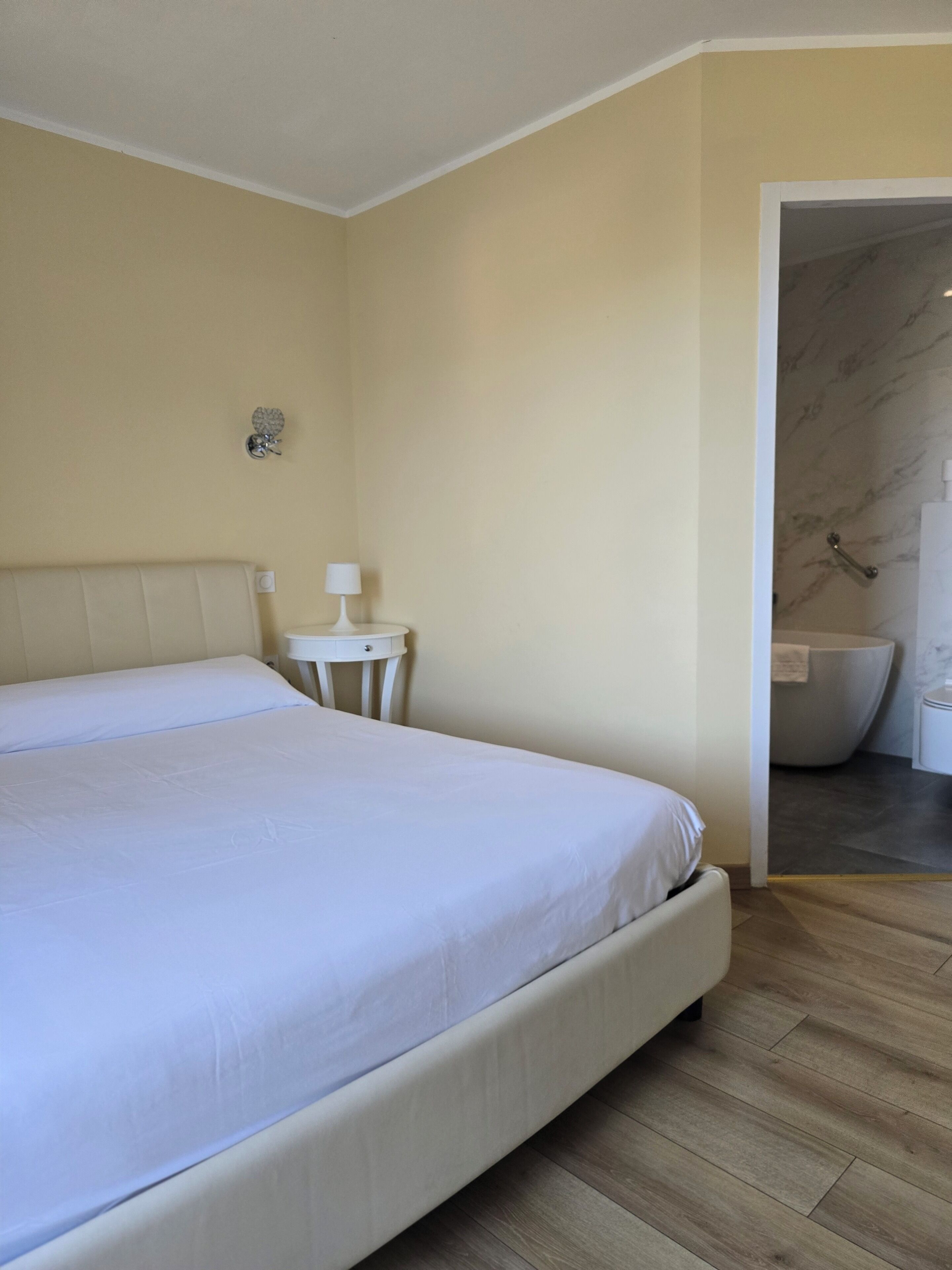 Un dormitorio con una cama, una mesita de noche con lámpara, y un baño visible a través de una puerta abierta.