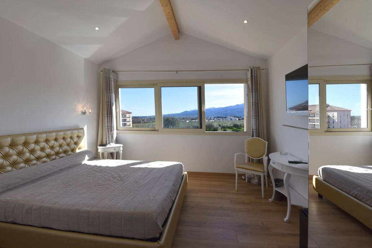 Une chambre avec un grand lit, un bureau et une chaise. Les fenêtres offrent une vue sur les montagnes et les bâtiments.