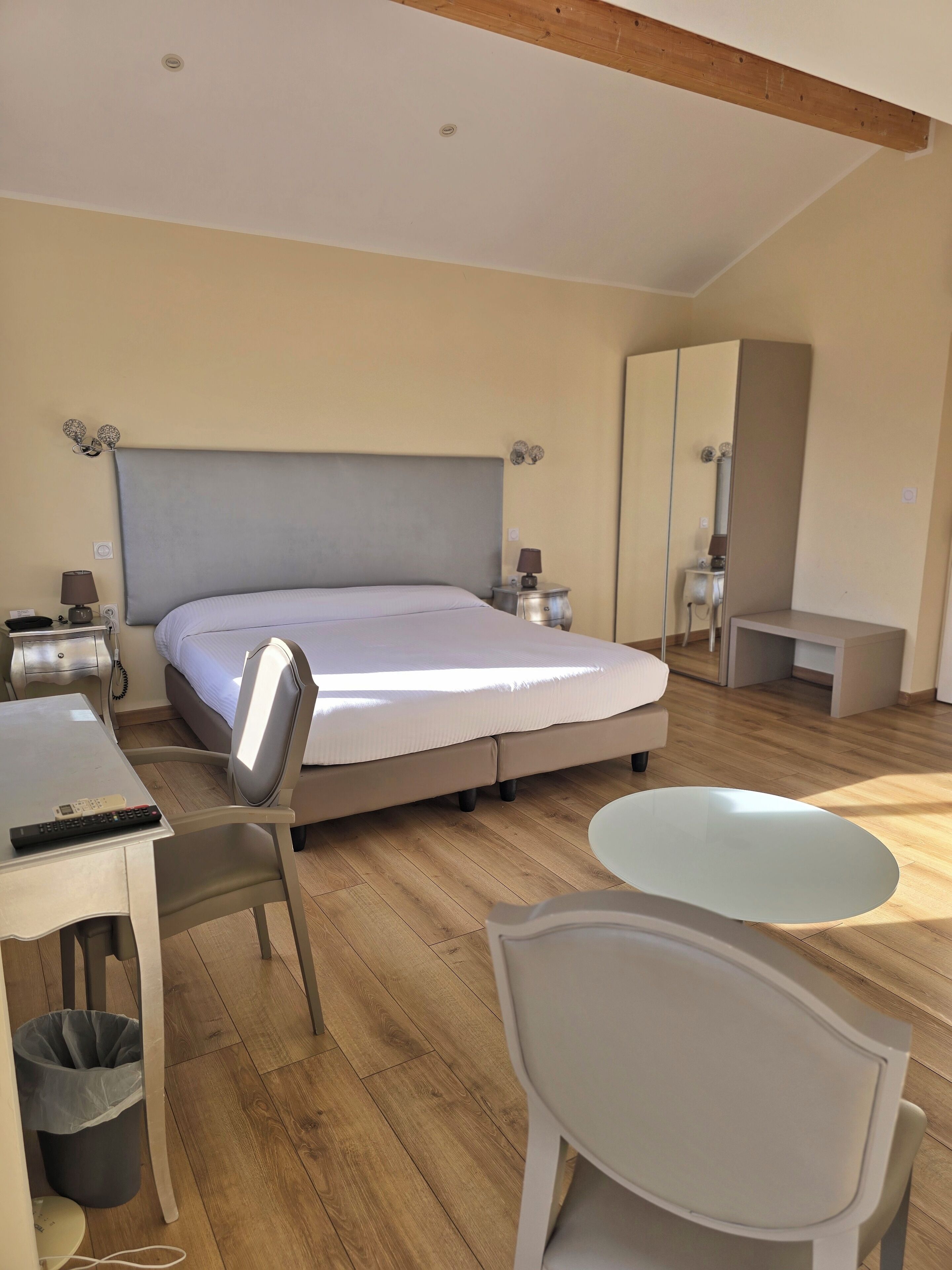 A modern hotel room with a large bed, a desk with a chair, a small table, and a trash bin.