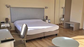 A modern hotel room with a large bed, a desk with a chair, a small table, and a trash bin.