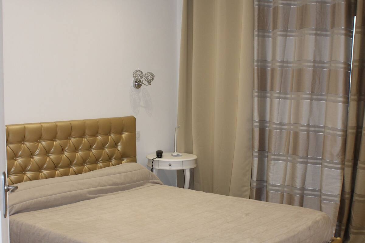 Une chambre à coucher avec un lit, une table de chevet, un luminaire fixé au mur et un rideau.