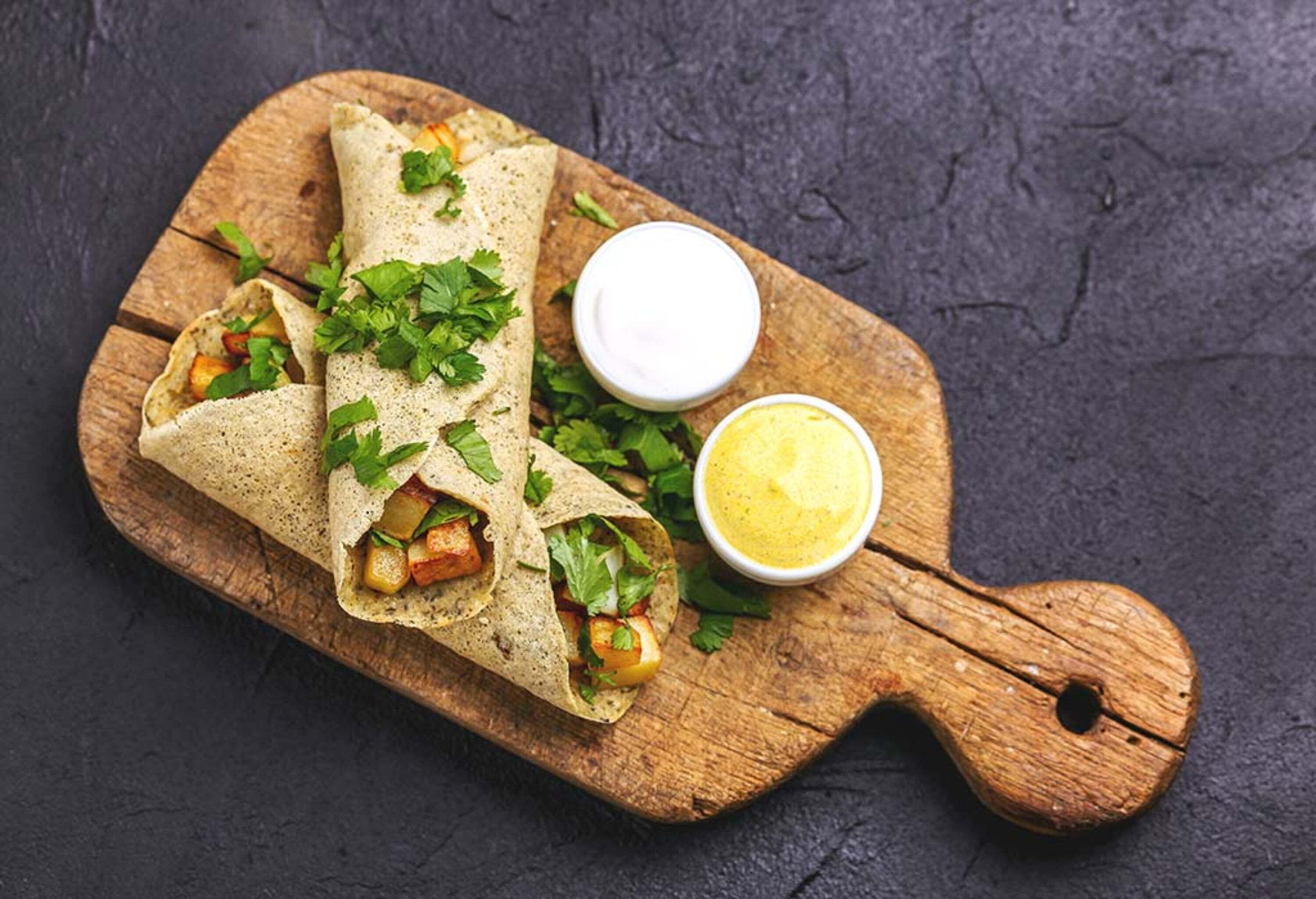A wooden board with three wraps containing vegetables and herbs, accompanied by two small bowls of sauce.