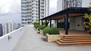 Una terraza en la azotea con cubierta de madera, macetas de piedra con plantas y vistas a edificios altos al fondo.