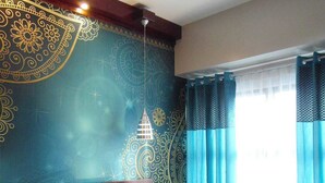 Una habitación de hotel con una cama grande, un cabecero con diseño decorativo, una mesita de noche y un ventanal con cortinas azules.
