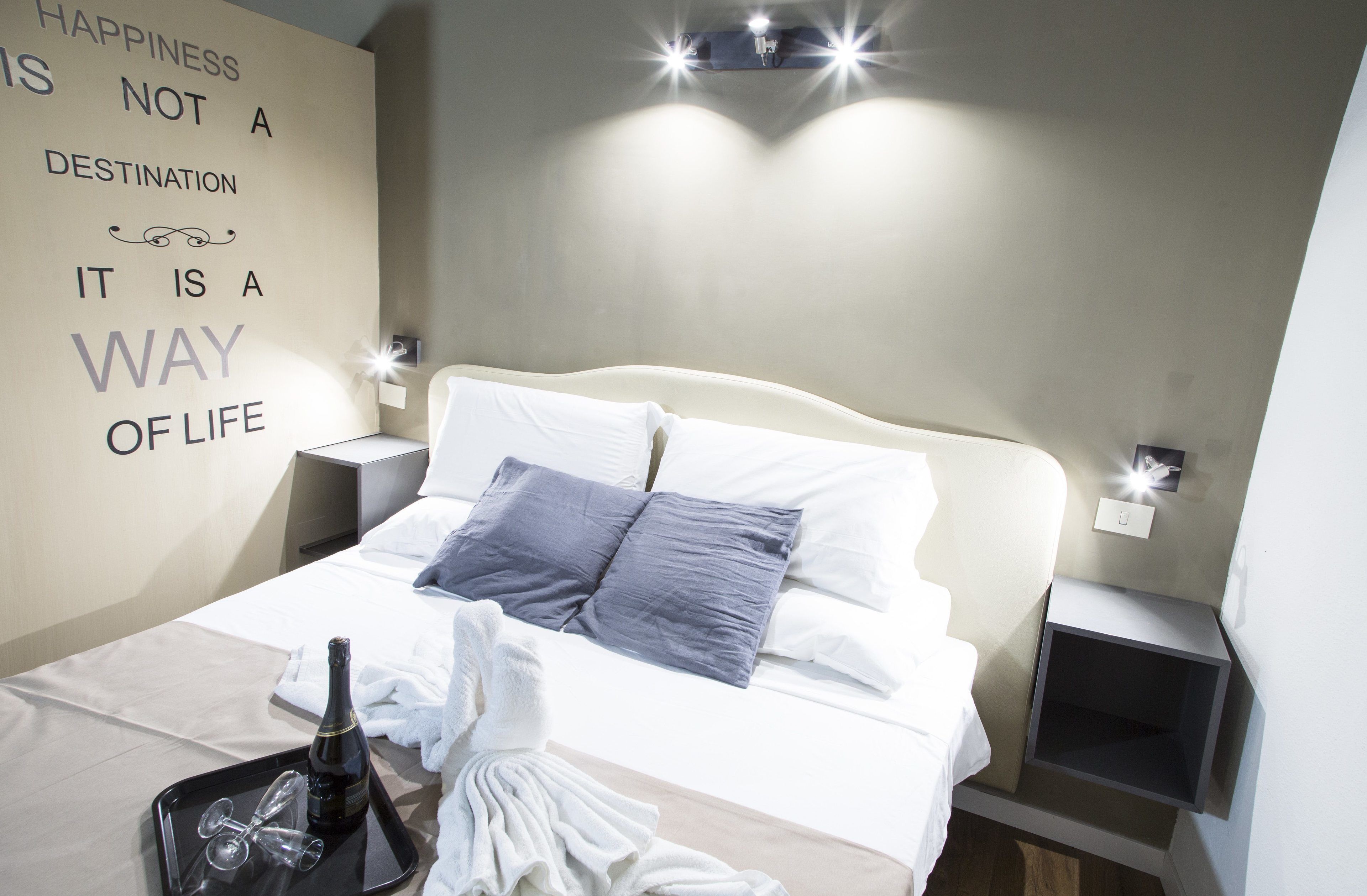 Une chambre d’hôtel avec un lit, des oreillers, un plateau avec une bouteille et des verres, et une citation inspirante affichée sur le mur.