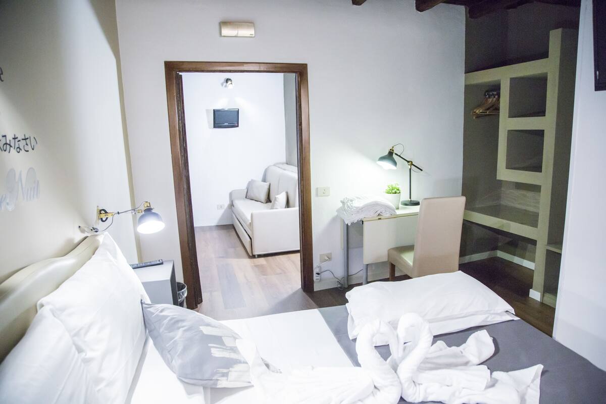 Une chambre d’hôtel avec un lit, une table de chevet, une lampe et une vue sur une autre chambre qui contient un canapé et un téléviseur.