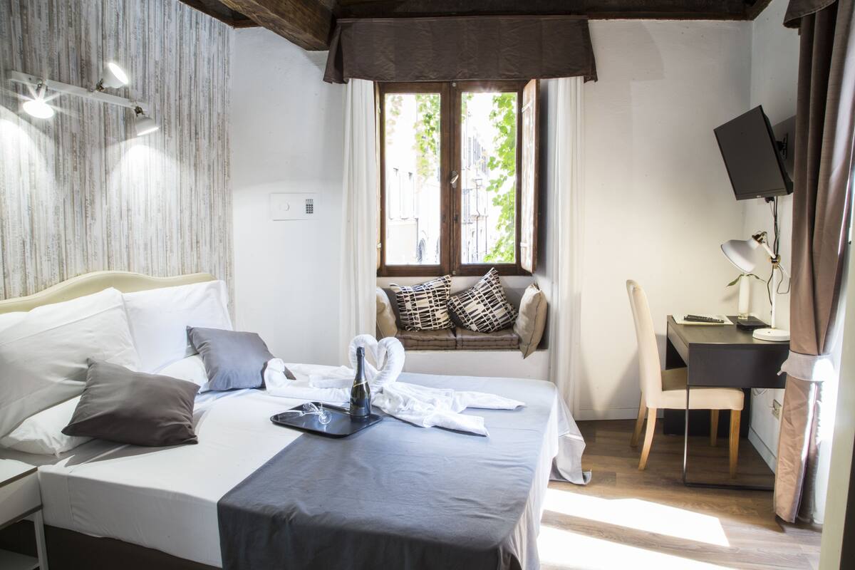 Une chambre d’hôtel avec un lit, un bureau, une chaise, une fenêtre avec des rideaux et un téléviseur fixé au mur.