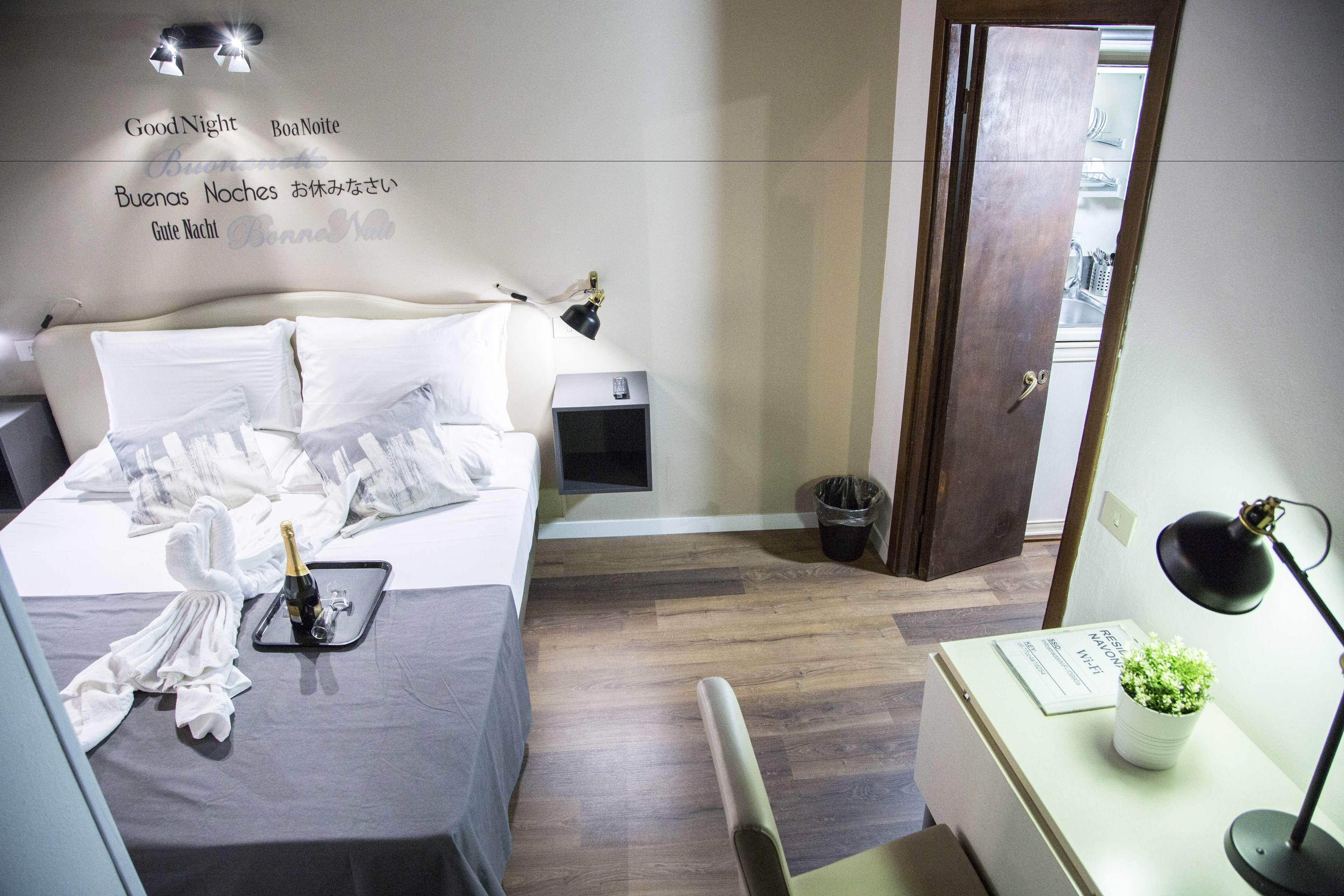 Una habitació d'hotel amb un llit ben fet, una taula de nit amb una làmpada, un petit safataire i una porta que dona a un bany.