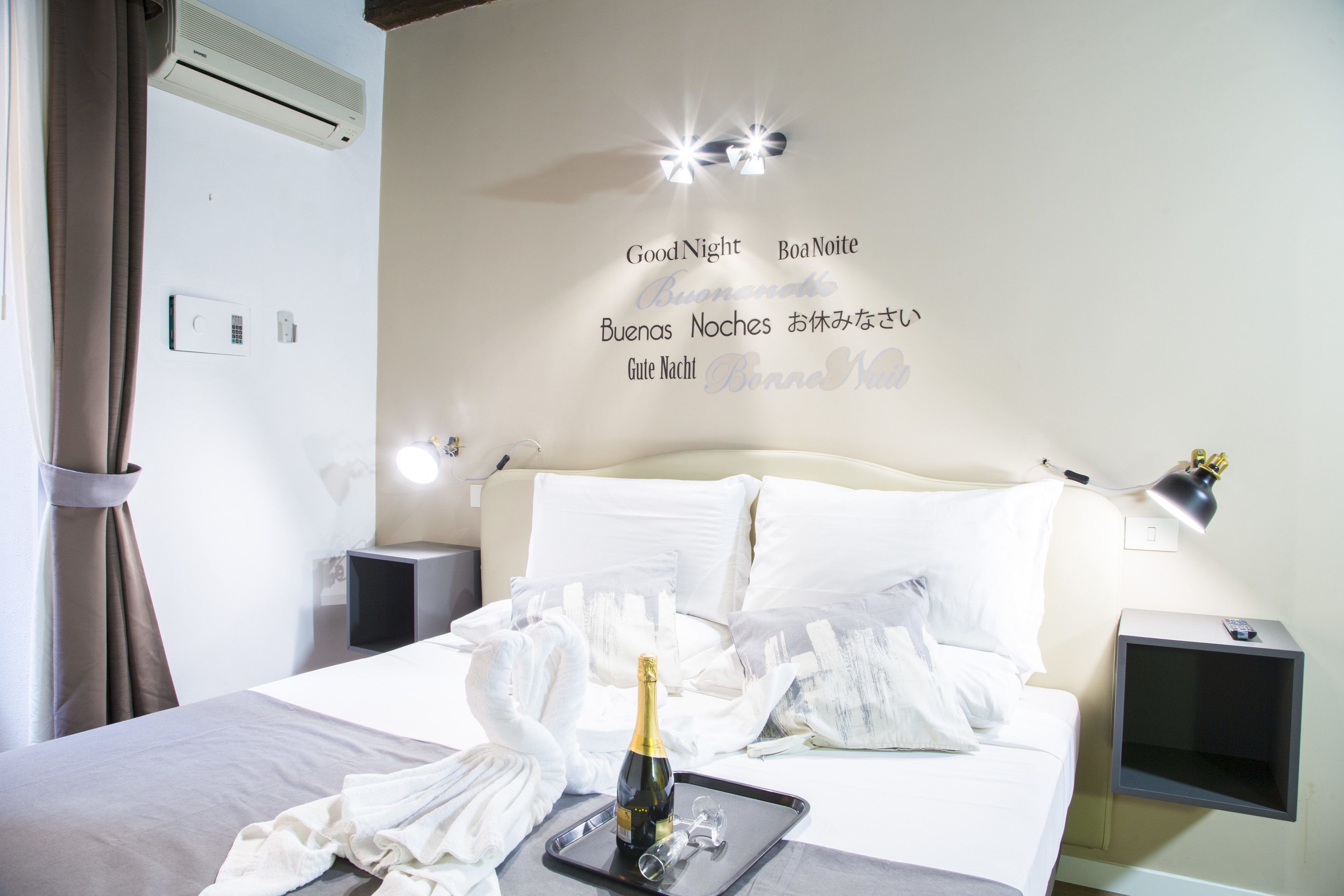A hotel room with a bed, a bedside table with a lamp, a wall clock, and a bottle of champagne on a tray.