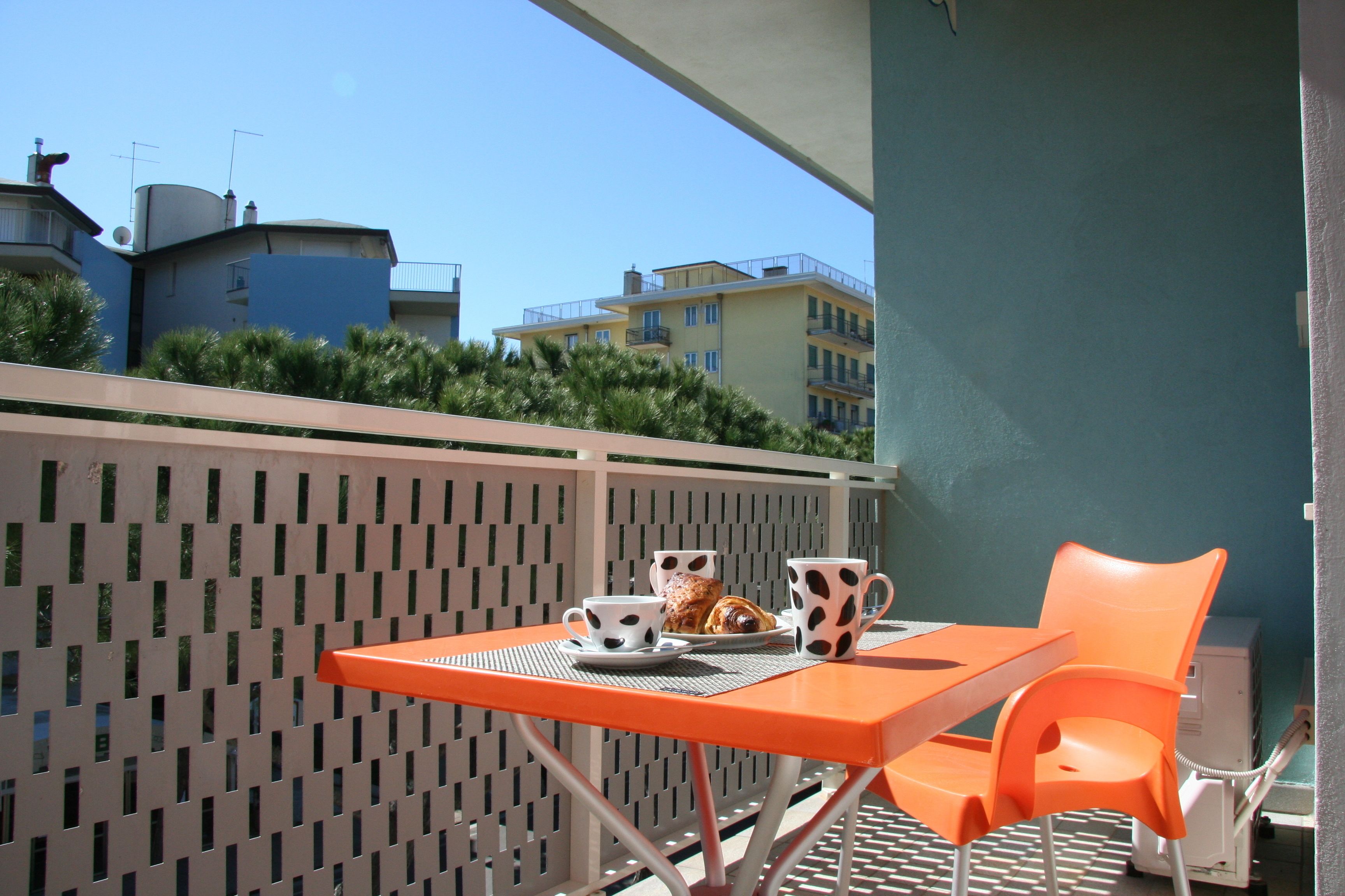 A balcony with a table and chairs, a small table with a cup and a pastry, and a view of buildings in the background.