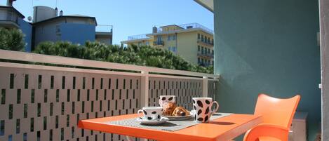 A balcony with a table and chairs, a small table with a cup and a pastry, and a view of buildings in the background.